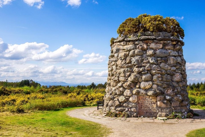 Stone cairn monument surrounded by grass and shrubs under a blue sky with clouds.