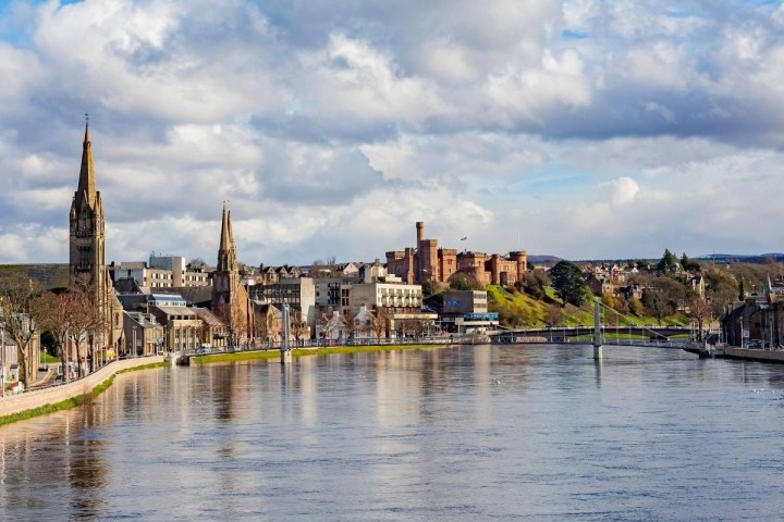 Riverside cityscape with clock towers and a distant castle under a partly cloudy sky.