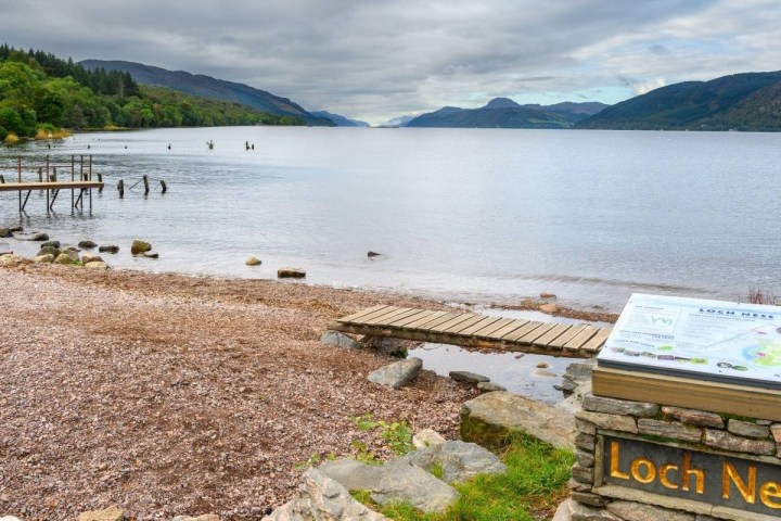 Loch Ness shoreline with a wooden dock and sign, surrounded by hills and cloudy sky.