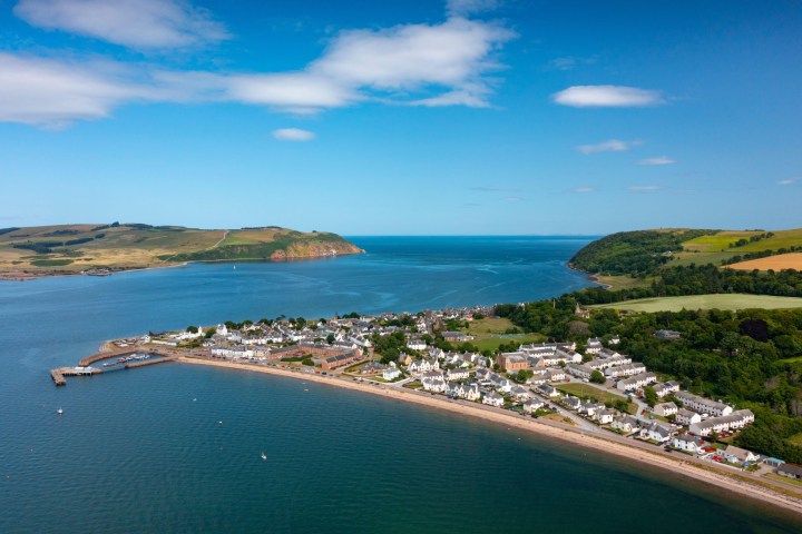 Coastal town with white houses, grassy hills, and blue sea under a clear sky.